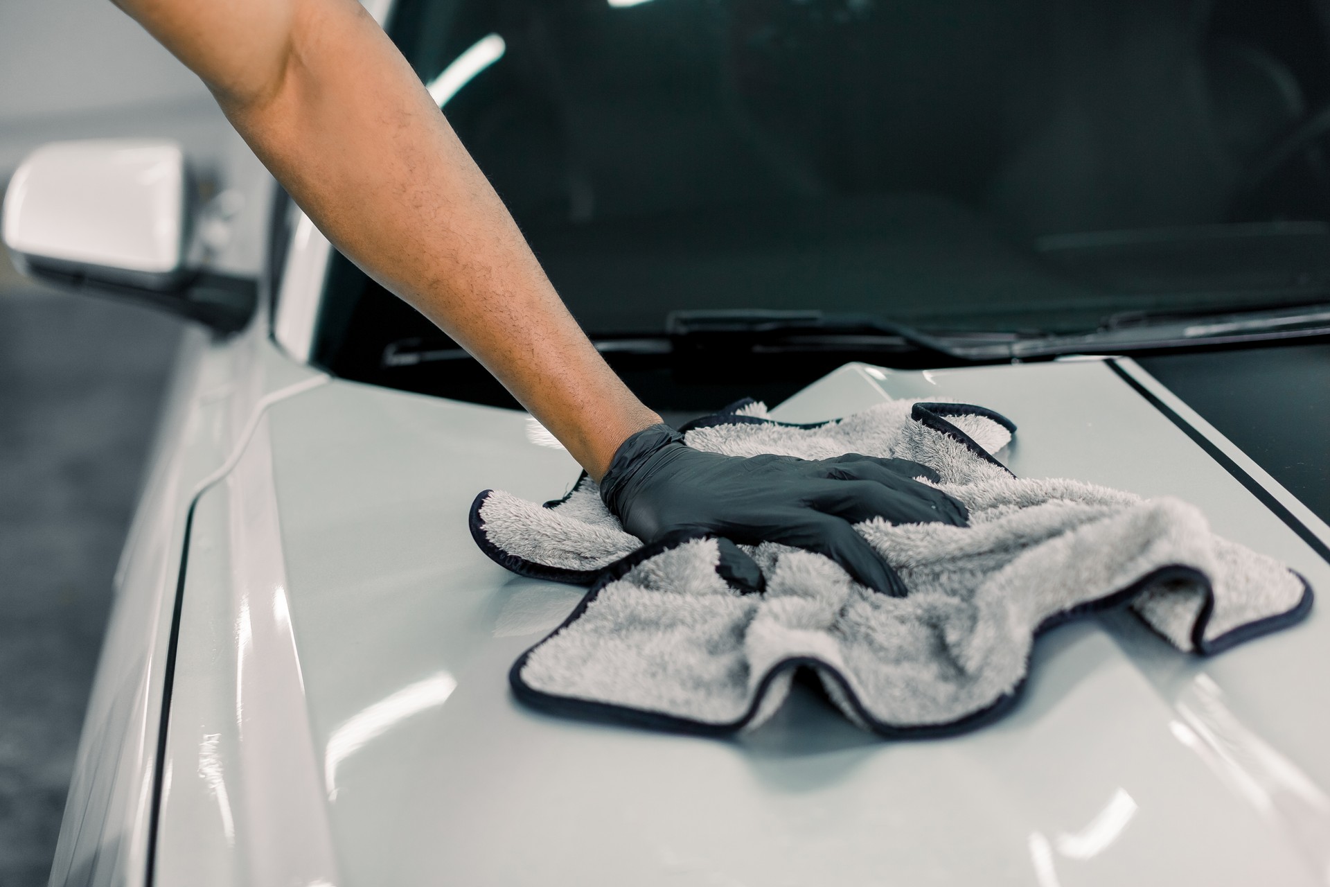 Car detailing, wash and cleaning concept. Cropped image of hand of male professional car wash worker in black rubber gloves, holding the gray microfiber and polishing the car hood of luxury white car